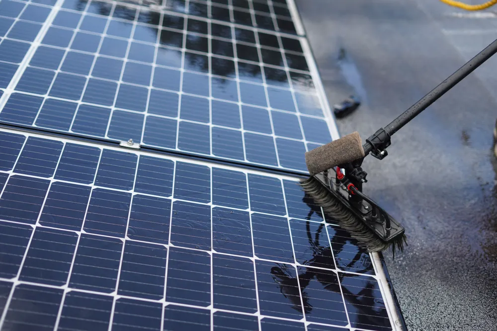 A soft-bristle brush on a water-fed pole cleaning a solar panel, water sheeting across the cells